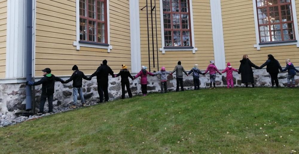 Lapsia ja aikuisia tekemässä ketjua Vesannon kirkon ympärille.
